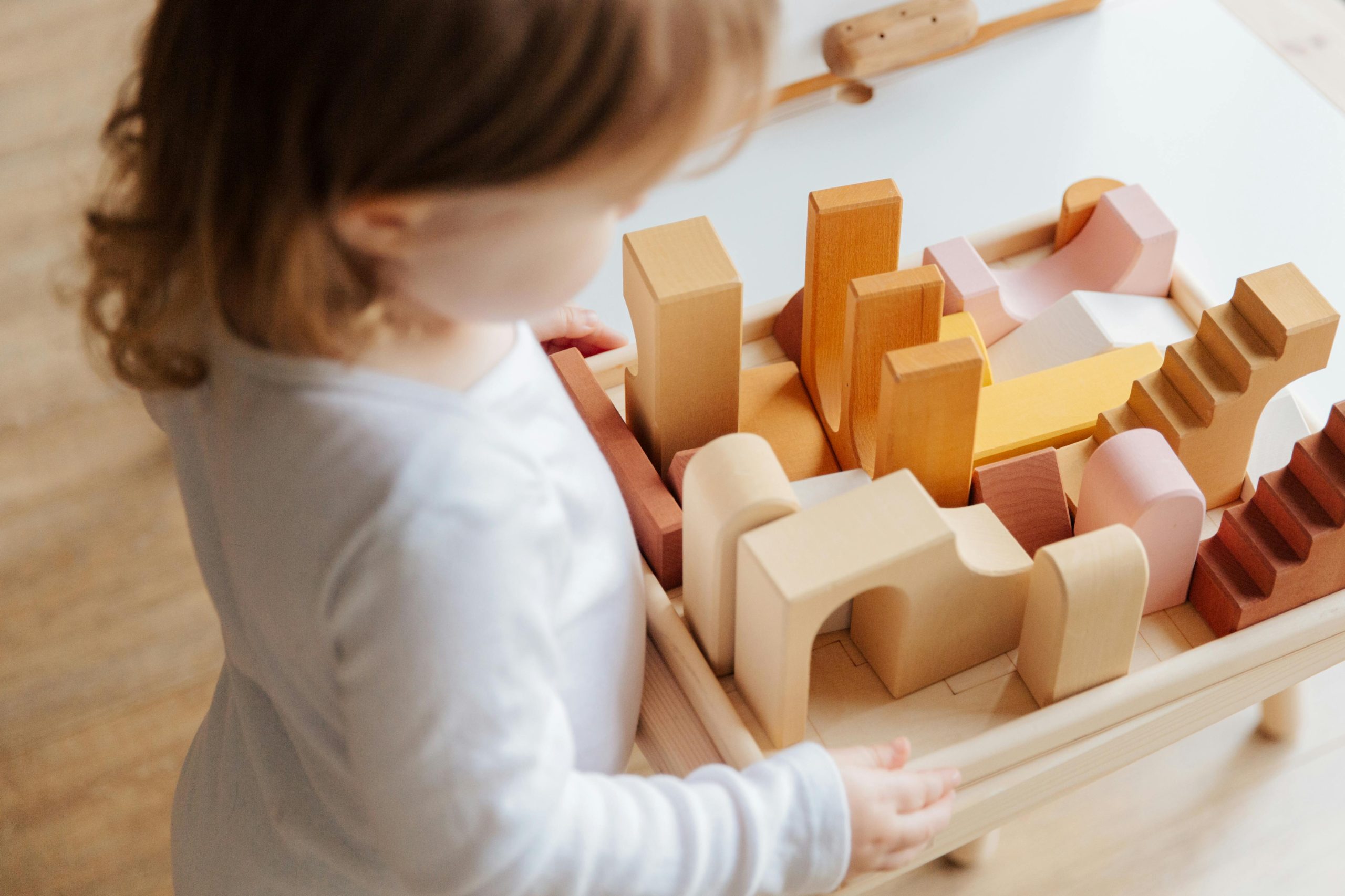 From above view of anonymous cute little girl in casual clothes standing near table and playing with wooden blocks at home